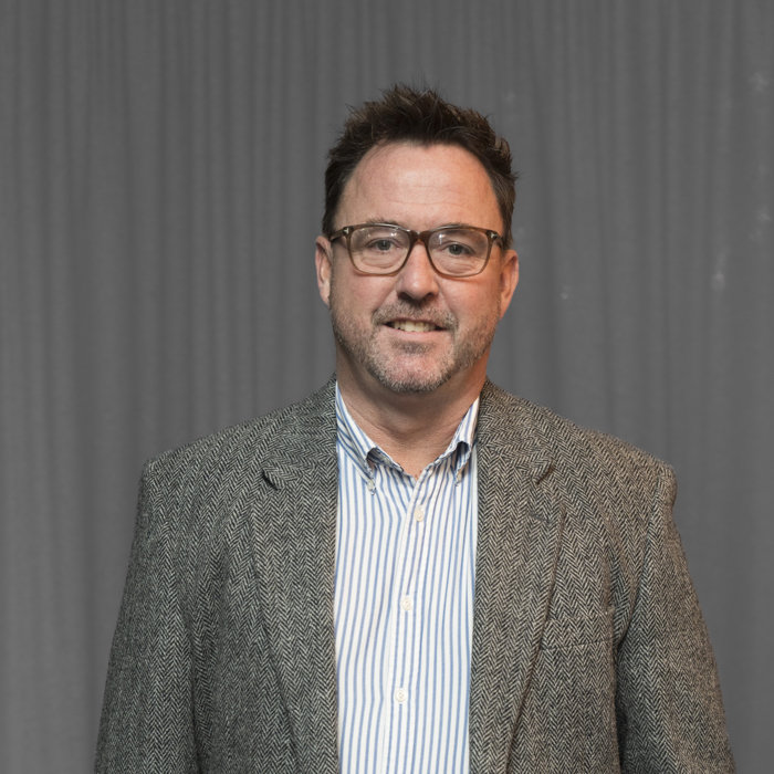 A man with short dark hair, glasses, and a beard is wearing a striped shirt and a tweed blazer, standing in front of a gray curtain.
