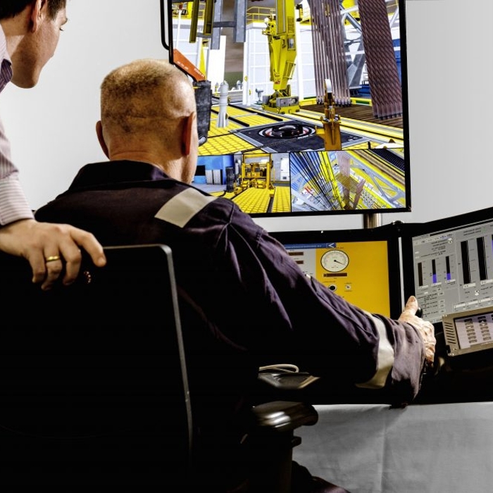 Two men in a control room monitor multiple screens displaying industrial machinery and data charts. One man sits while the other leans over, observing the screens.