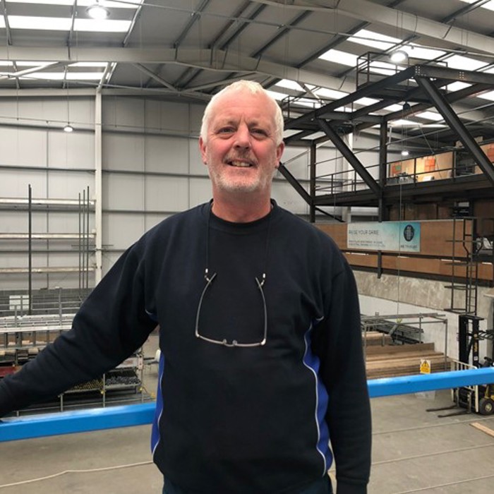 A man with a grey beard and a dark sweater stands inside an industrial warehouse, smiling at the camera.