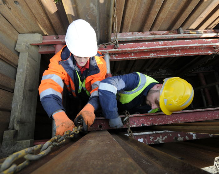Two construction workers in safety gear are handling a metal chain inside a trench with surrounding wooden and metal supports, skillfully applying their training to ensure a safe and efficient process.