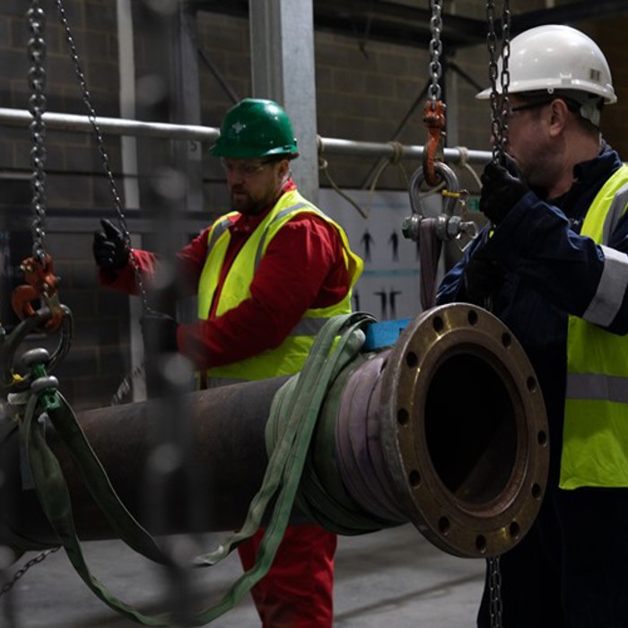 Two workers in safety vests and helmets lift a large pipe using a chain hoist in an industrial setting.