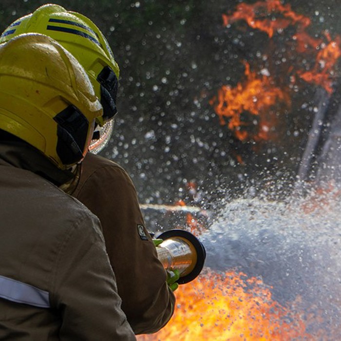 Two firefighters in yellow helmets spraying water at an intense fire with visible flames.