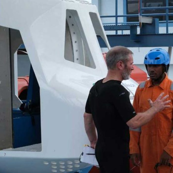 A person in a black shirt instructs two individuals in orange uniforms and blue helmets standing next to a large white apparatus.