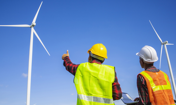 Two workers in safety gear stand outdoors, observing wind turbines against a clear blue sky. One points upward while the other holds a laptop, highlighting the importance of ongoing training in mastering these renewable energy systems.