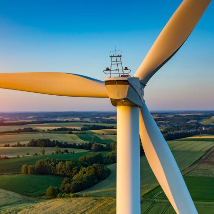 Close-up view of a wind turbine with a scenic countryside landscape and a setting sun in the background. Fields and hills are visible under a clear sky.