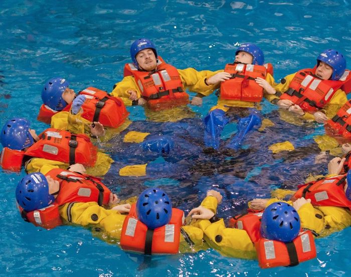 A group of people wearing orange life jackets and helmets form a circle while floating in a pool, participating in a training exercise.