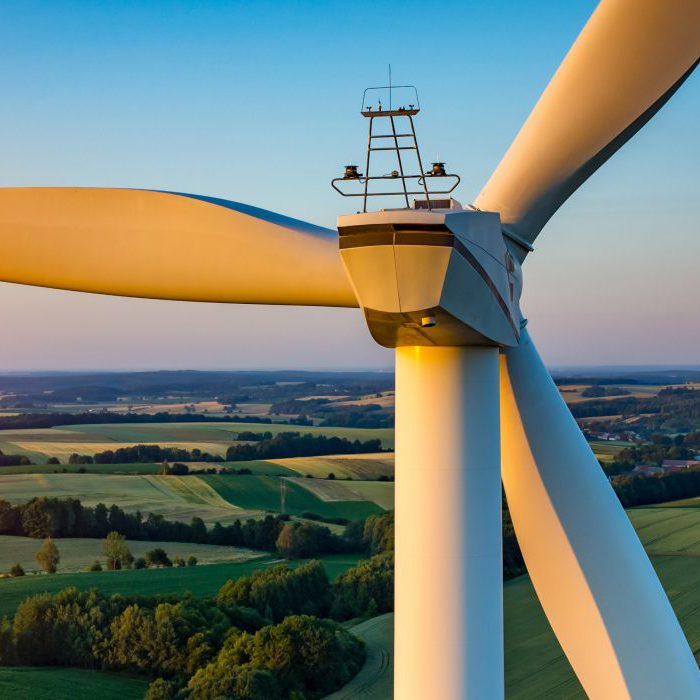 Close-up of a wind turbine with three blades, set against a backdrop of sprawling green fields and a setting sun. The sky is clear with a gradient from blue to orange near the horizon.