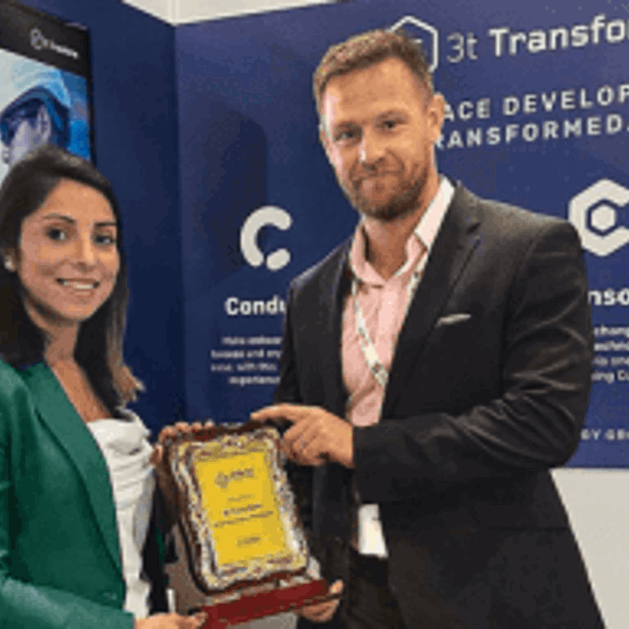 Two people stand in front of a booth backdrop displaying "3t Transform". The woman in a green blazer is holding an award, while the man in a suit poses beside her.
