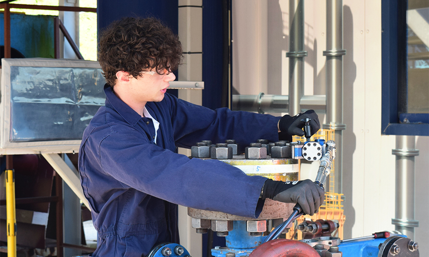 A person in a blue jumpsuit operates industrial equipment, adjusting controls on a blue metal framework in a facility with pipes and machinery.