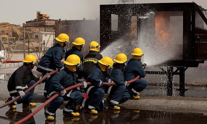 A group of firefighters in yellow helmets and dark uniforms spray water at a burning structure during a training exercise.