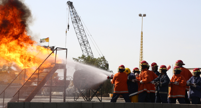 Firefighters in orange uniforms and red helmets use a hose to spray water on a large industrial fire with thick smoke. A crane and flag are visible in the background.