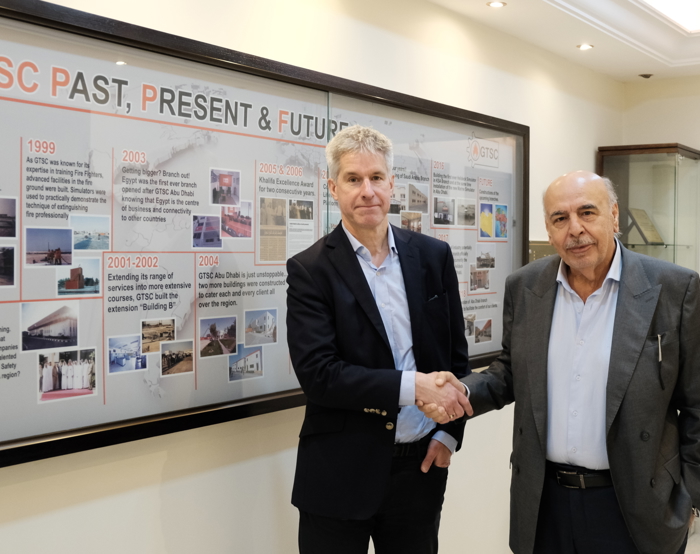 Two men shaking hands in front of a timeline display board labeled "GTSC Past, Present & Future" in an office, setting the stage for future training initiatives.