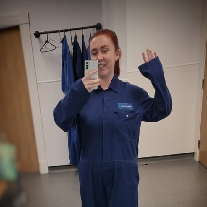 A person with red hair is taking a mirror selfie in a room, wearing a blue jumpsuit and holding a smartphone. There are hanging blue jumpsuits in the background.