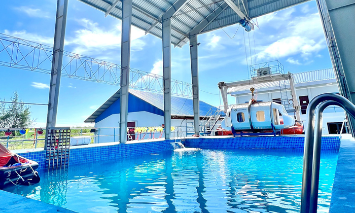 An indoor pool with a metallic roof structure overhead. A large piece of equipment is positioned beside the pool, and a blue-roofed building is visible in the background.