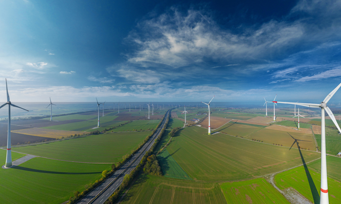 Aerial view of a wind farm with multiple wind turbines spread across a landscape of green fields and a road running through the middle under a partly cloudy sky, showcasing the transformation from military to wind energy initiatives.