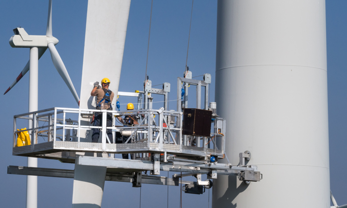 Two workers in safety gear stand on a suspended platform performing maintenance on a large wind turbine, showcasing how skill sets like those from military to wind energy can be seamlessly transferred, all against a clear blue sky.